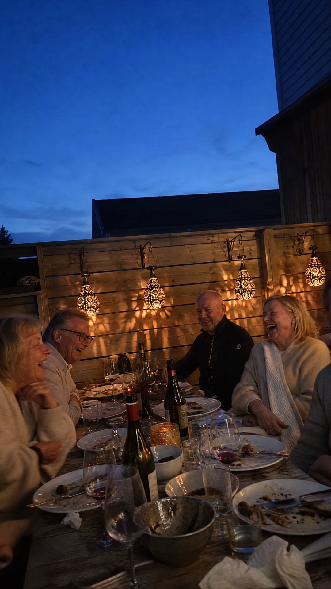 Friends laughing around a dinner table on a deck at dusk, with Light Drop lanterns glowing along the fence